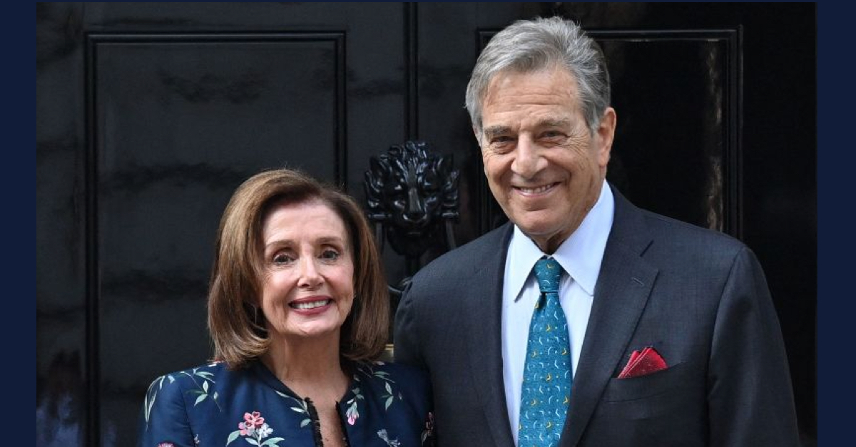 Nancy Pelosi is wearing a dark blue jacket with a design of pink flowers. Paul Pelosi is wearing a dark suit with a blue tie, white shirt, and red pocket square. They are standing in front of 10 Downing Street in London.
