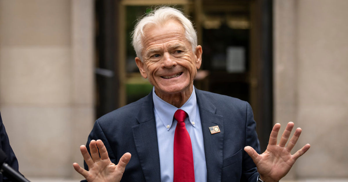 Peter Navarro is seen with his hands raised, palms toward the camera. He appears to be speaking. He is wearing a dark blue jacket, light blue shirt, and red tie, and on his lapel an American flag pin.