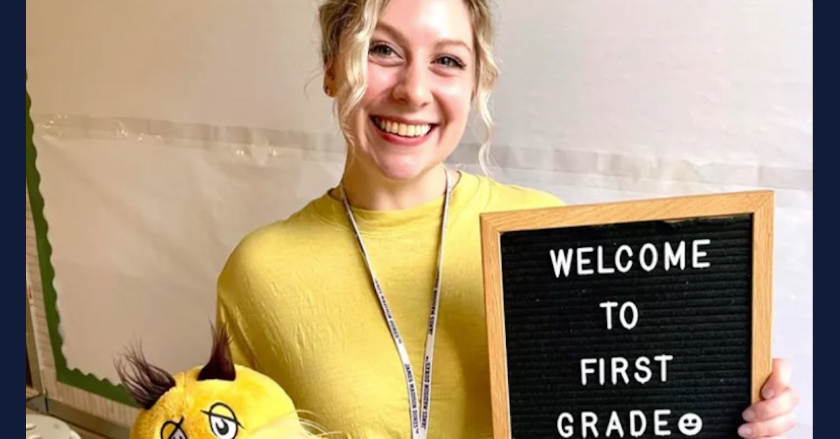 Abby Zwerner is wearing a yellow sweater and holding a sign that says "Welcome to First Grade." She is smiling and looking directly at the camera.