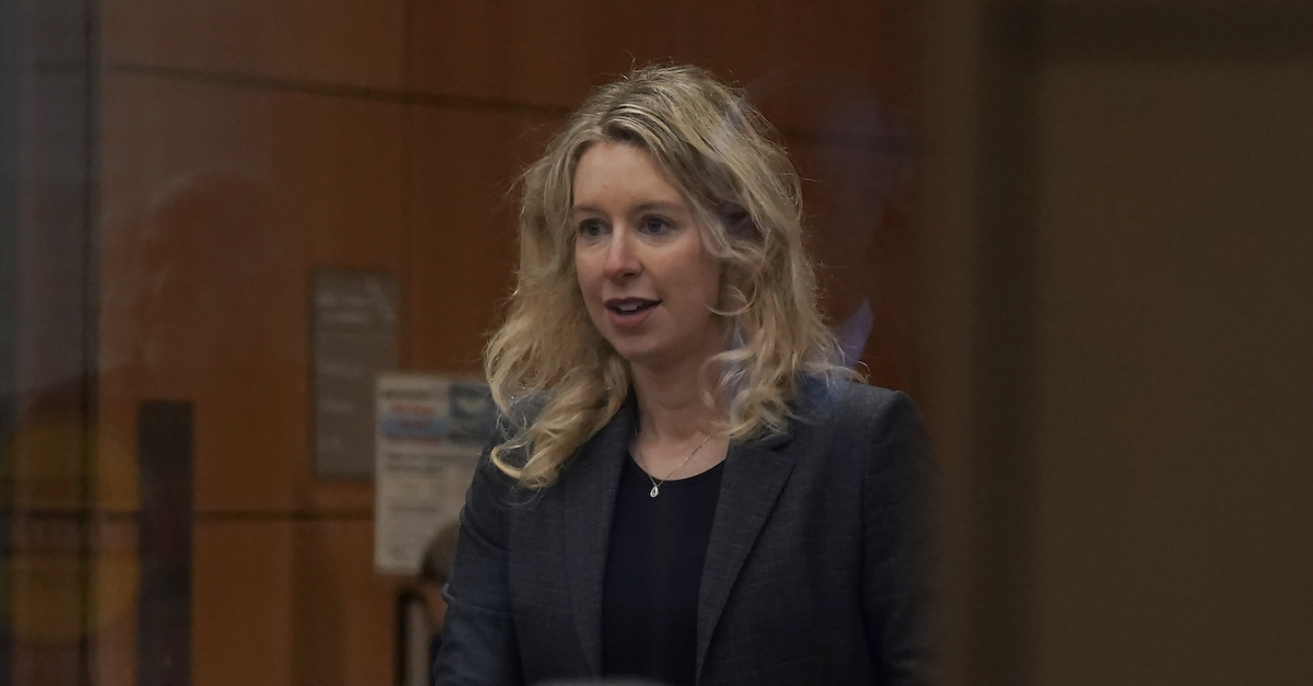 Elizabeth Holmes has blonde hair and is wearing a dark blue suit over a black shirt. She is standing behind a podium and partitions inside a wood-paneled courtroom and appears to be speaking to someone off-camera.