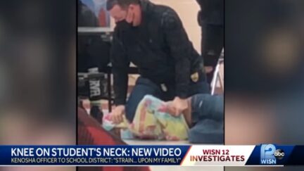 Kenosha police officer Shawn Guetschow is seen placing his right knee on the neck of a student he has pinned to the ground. The student is lying on her side and is wearing a tie-dyed sweatshirt.